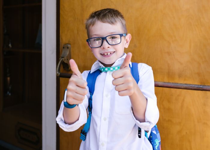 A cheerful boy with glasses and a backpack giving a thumbs up outside school