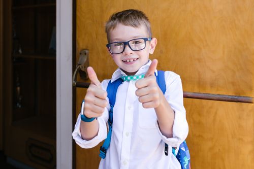 Free Cheerful boy wearing glasses with a backpack gives thumbs up outside school entrance. Stock Photo