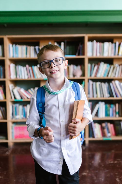Free A cheerful boy in a school uniform stands in a library holding books, ready for school. Stock Photo