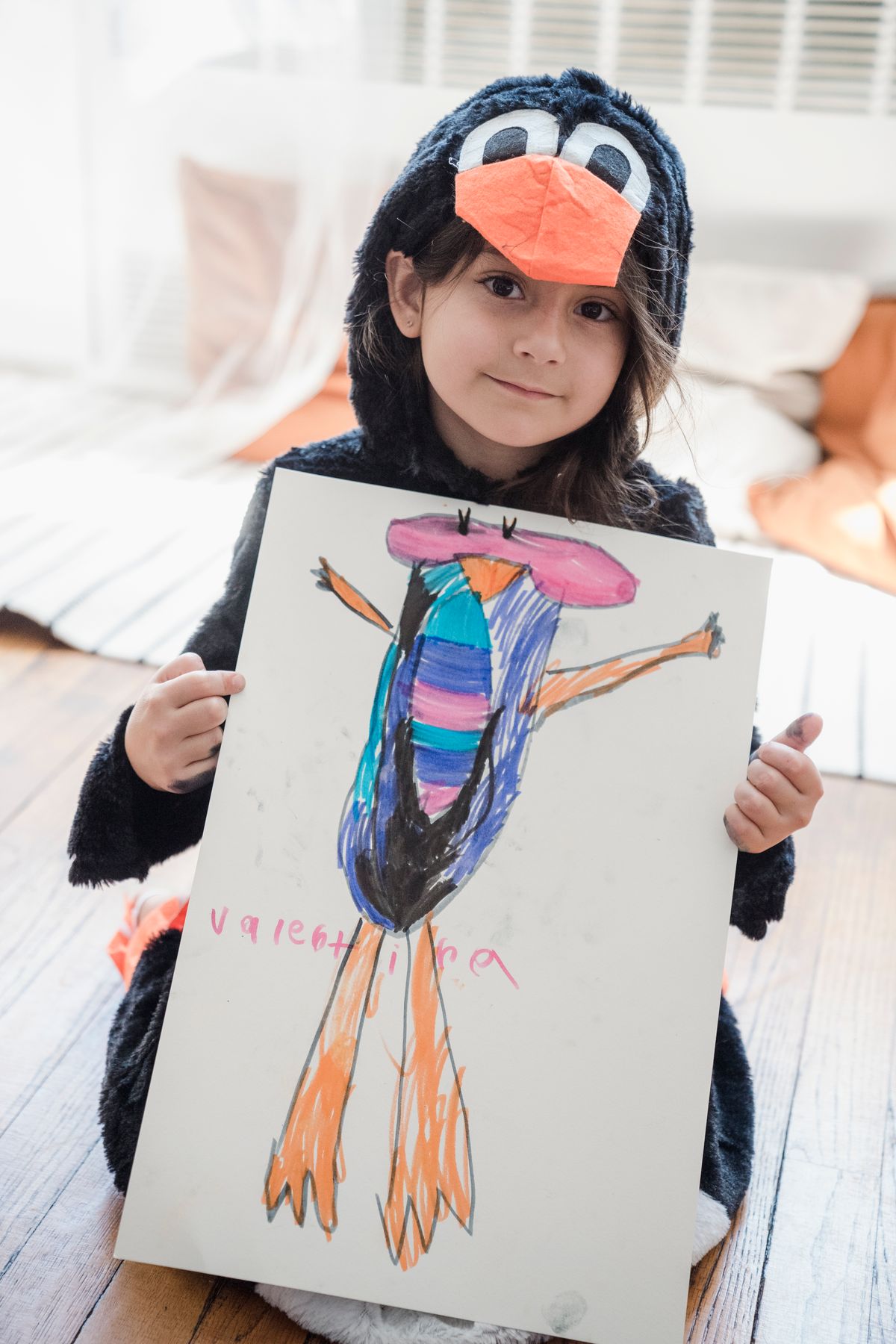 Young girl dressed as a penguin proudly displaying her whale artwork indoors