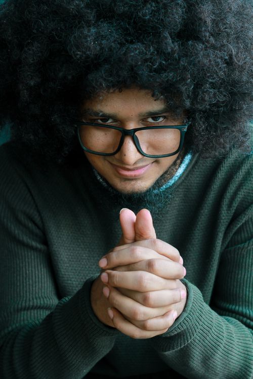 Free Portrait of a young man with afro hair and glasses smiling with hands clasped. Casual and approachable. Stock Photo
