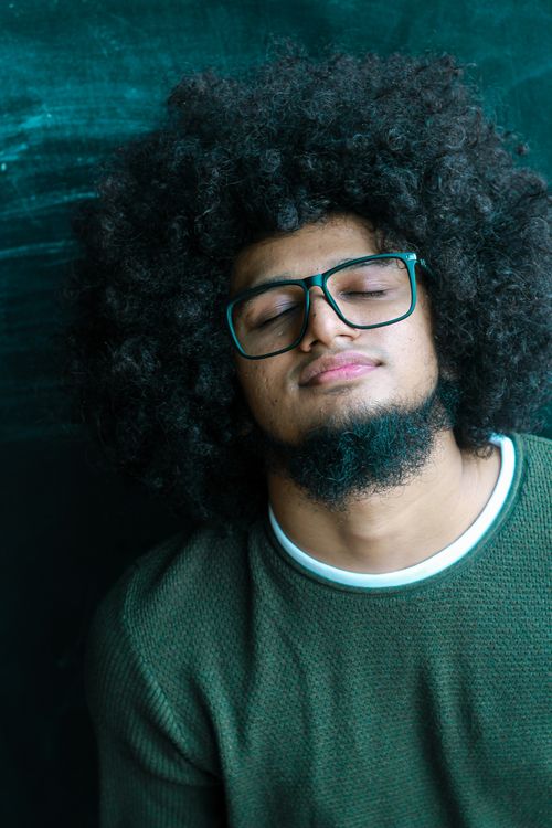 Free A young man with afro hair and glasses resting against a teal background. Stock Photo