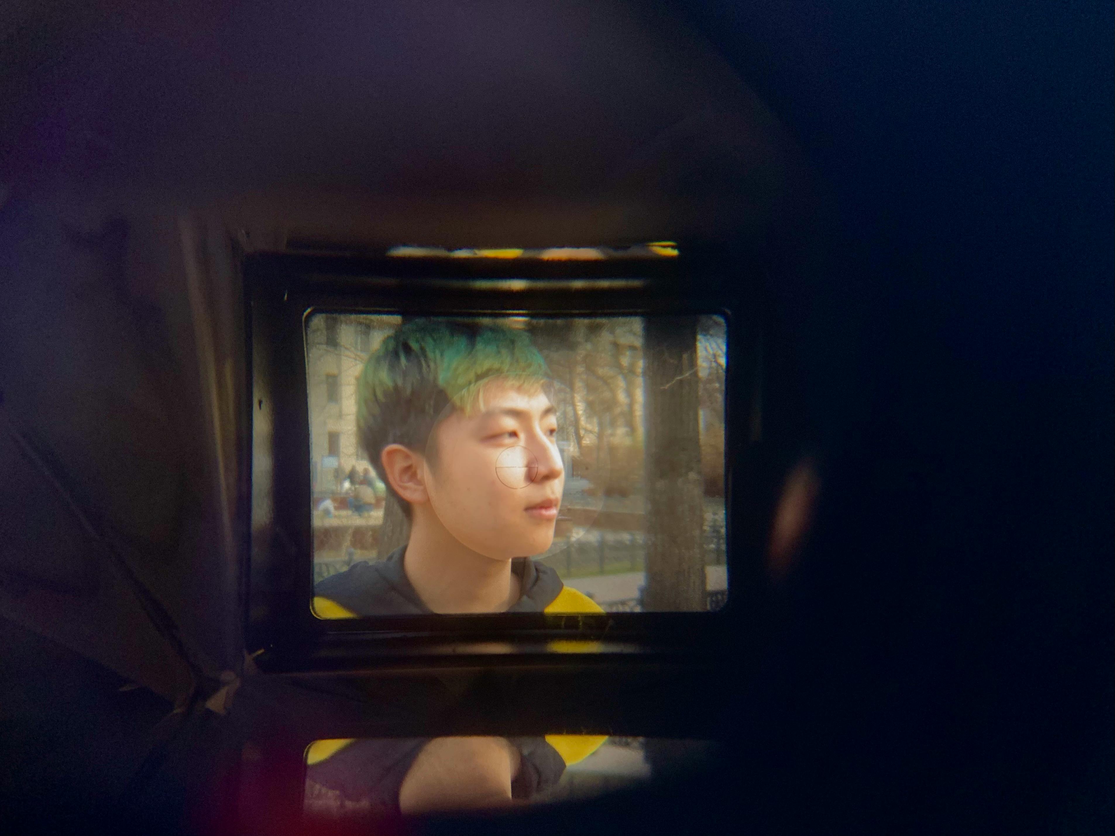 Creative portrait of a teenage boy with green hair seen through a camera viewfinder outdoors.