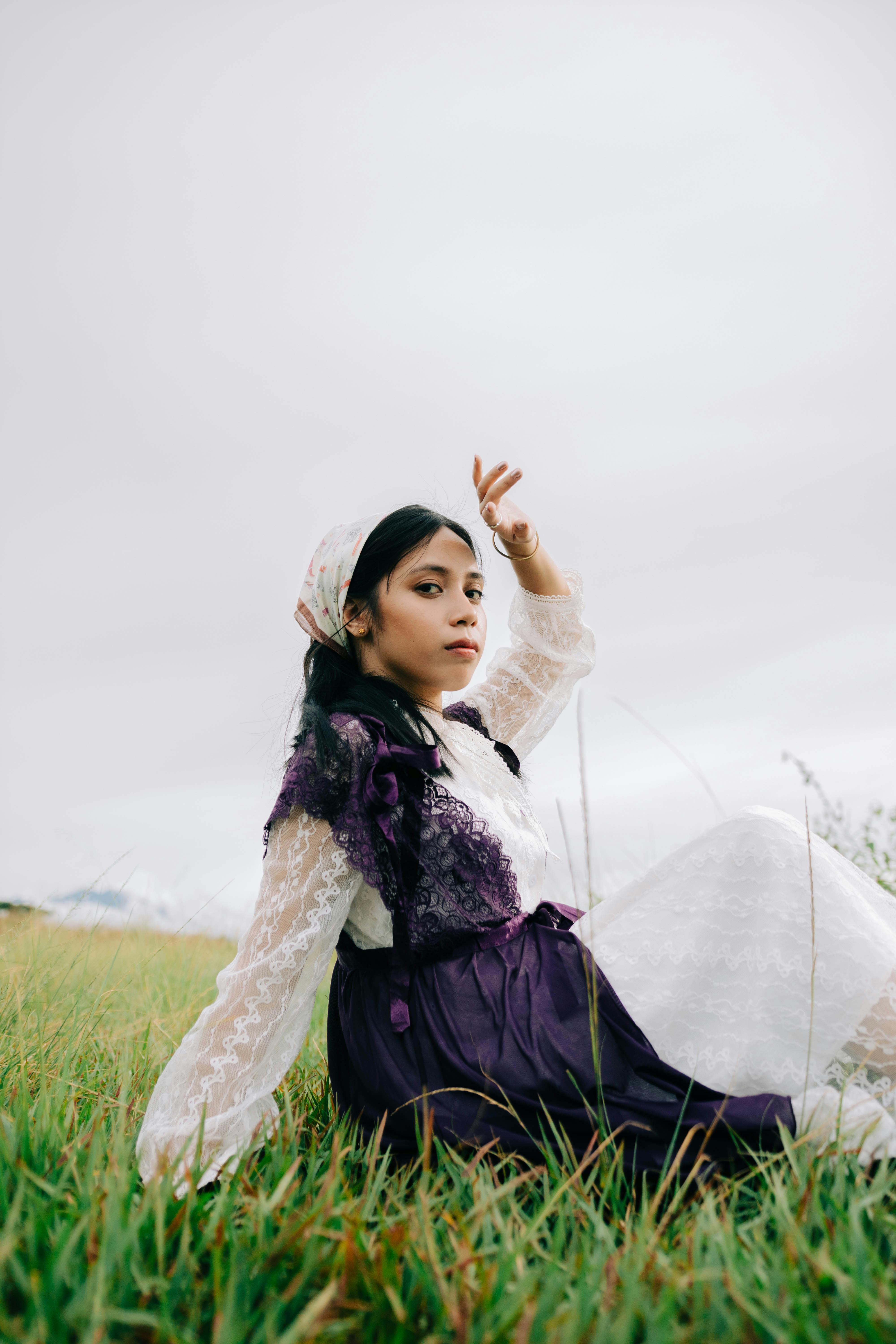 Woman in a white and purple dress sitting gracefully on a grass field outdoors.