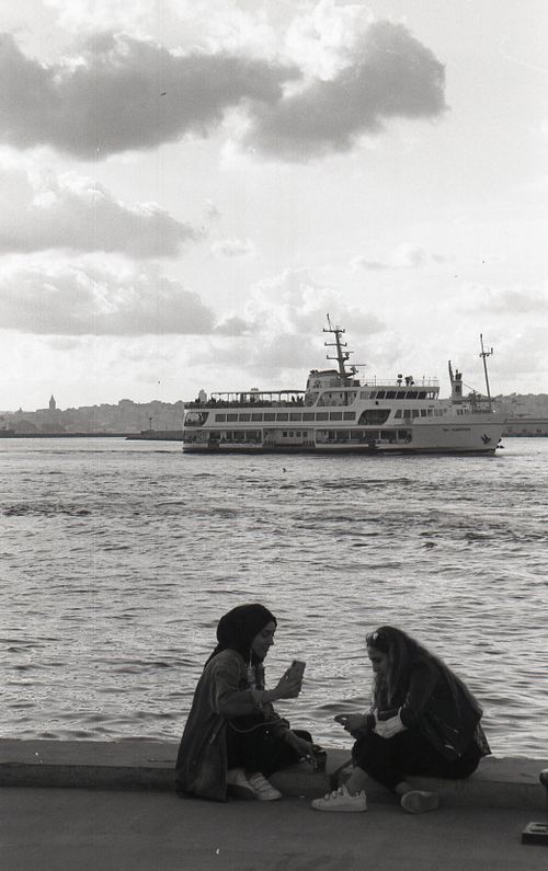 Free Black and white image of women sitting by the waterfront, with a ferry in the background. Stock Photo