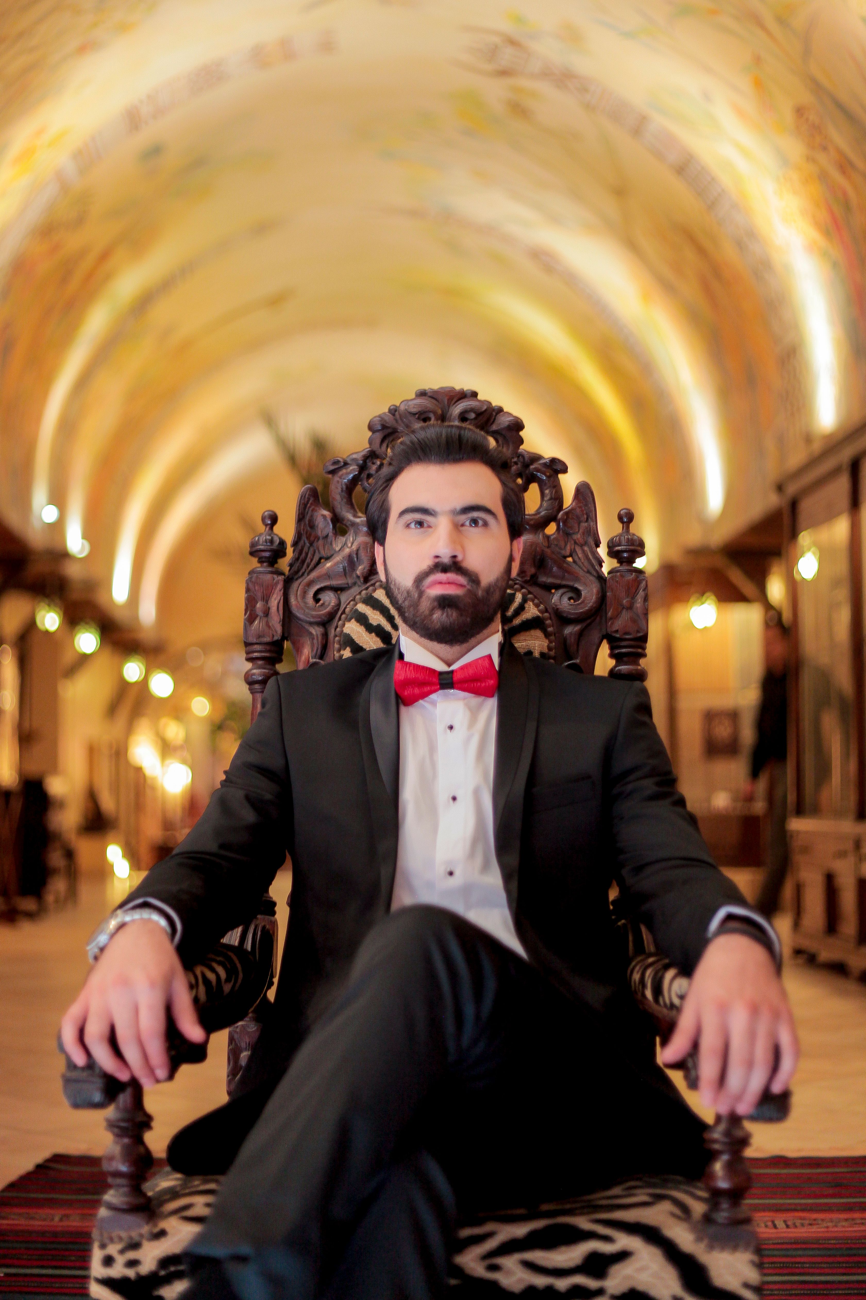 Sophisticated man in black suit and red bow tie seated in an ornate armchair in ornate hallway.