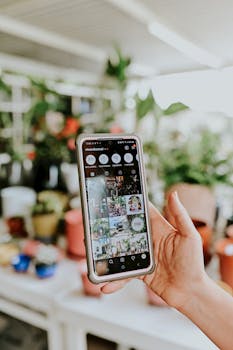 Hand holding smartphone with Instagram app open, blurred background of plants.