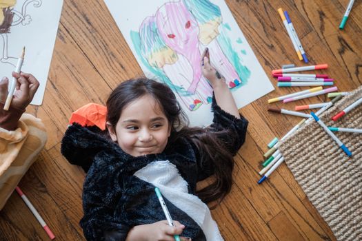 A child in an animal onesie drawing colorful pictures on a wooden floor, enjoying creative playtime.