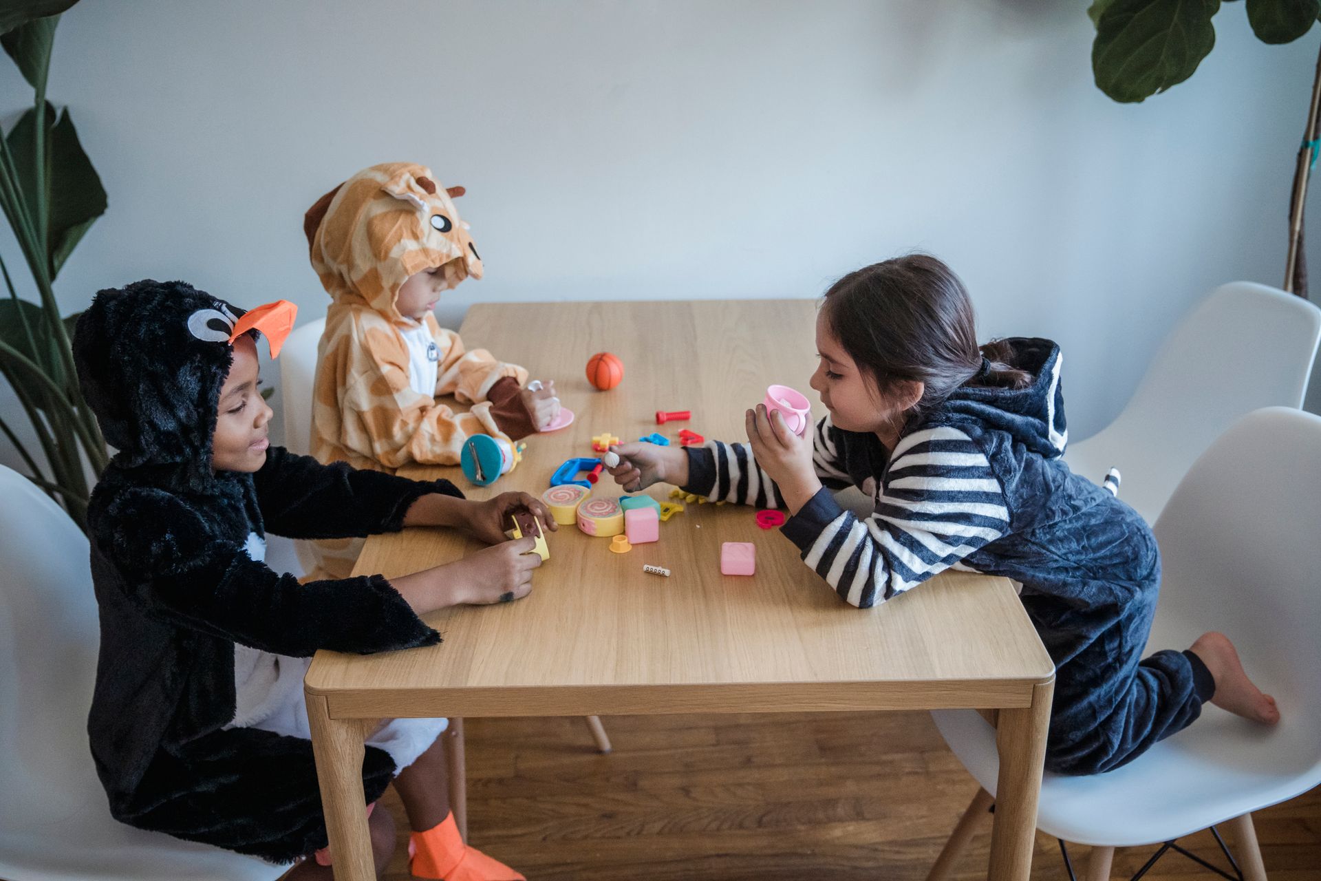 Three children wearing animal costumes play with colorful toys at a wooden table indoors.