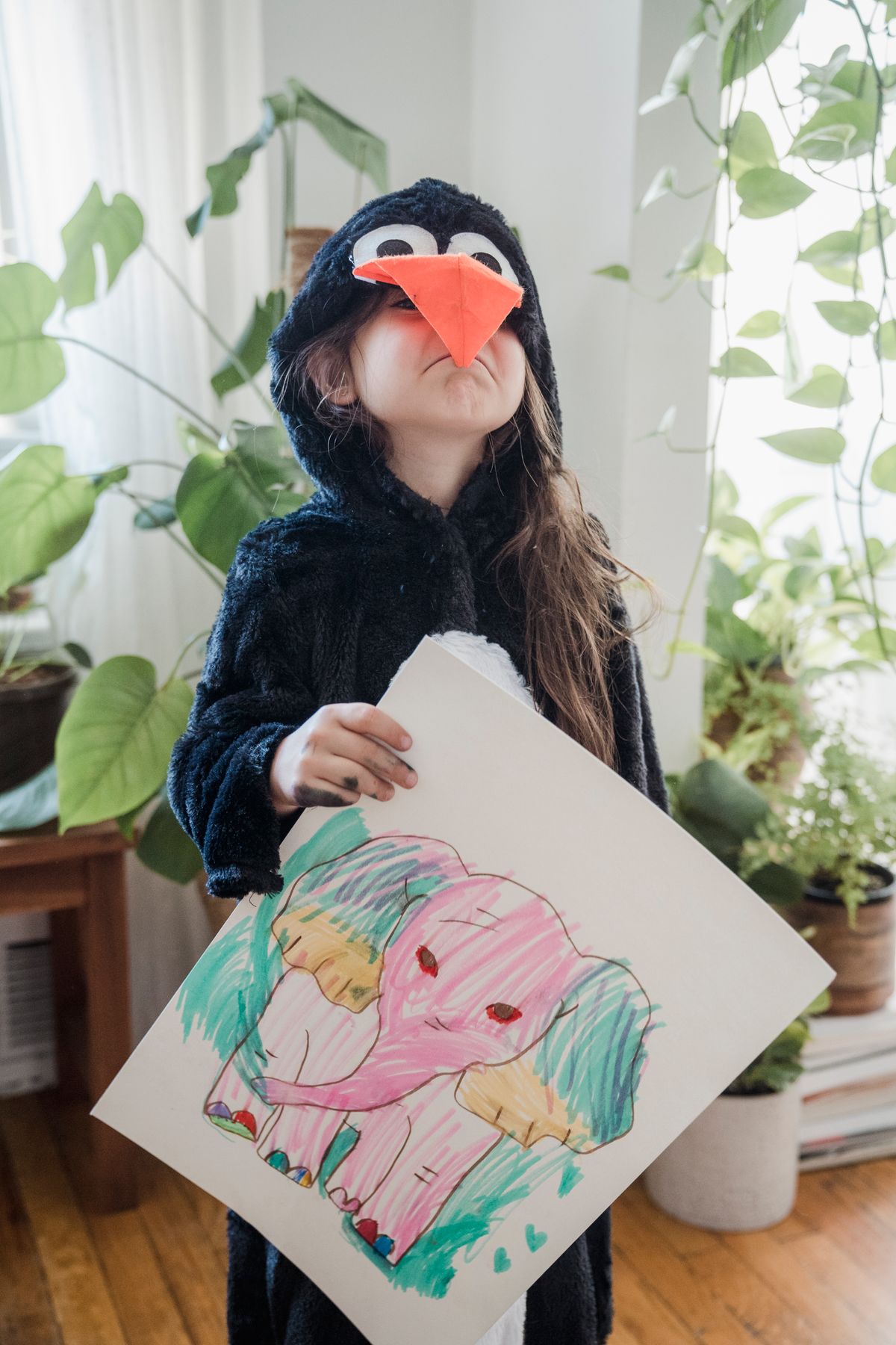 Child in a penguin costume proudly holding up a colorful elephant drawing