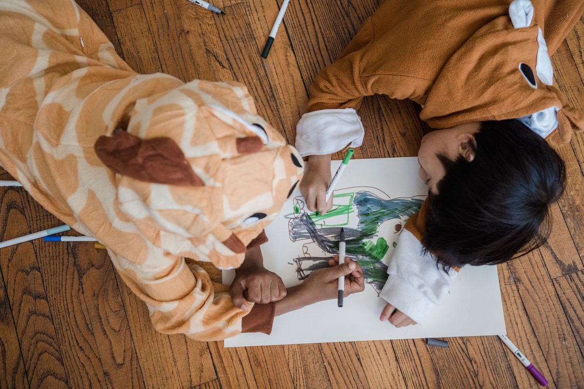 Two kids in animal costumes drawing a hedgehog with markers on a wooden floor