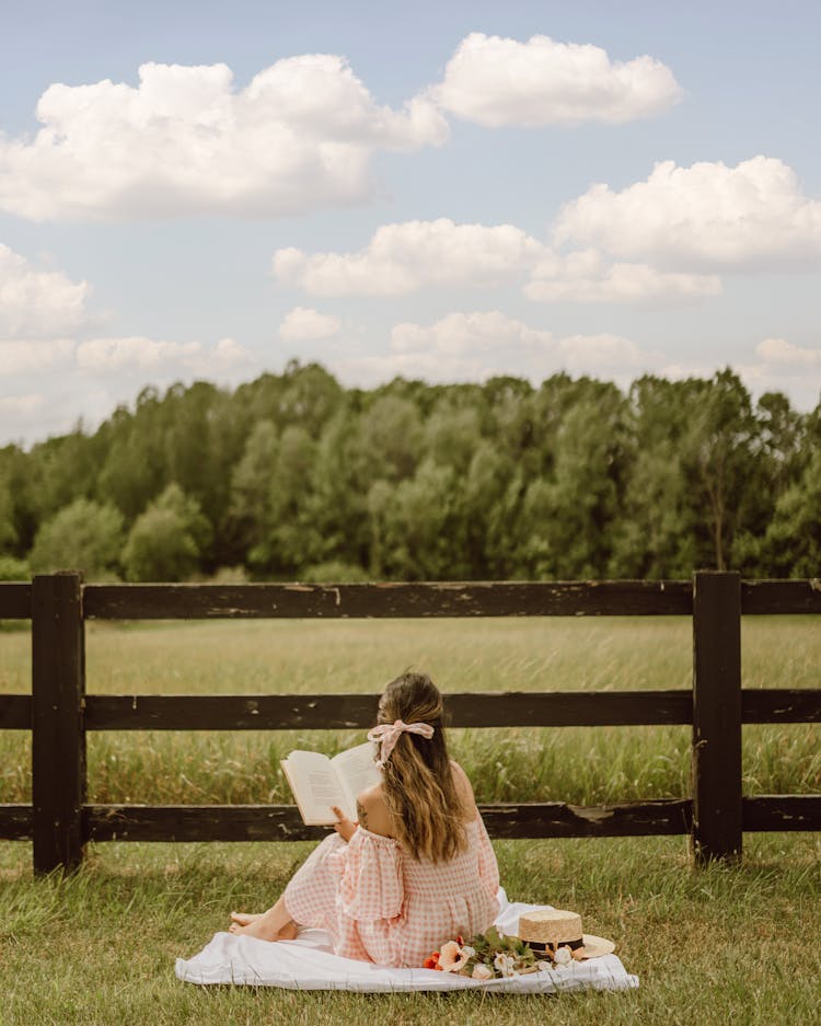 Back View Of A Woman Reading A Book While Sitting On A Picnic Blanket Near The Wooden Fence