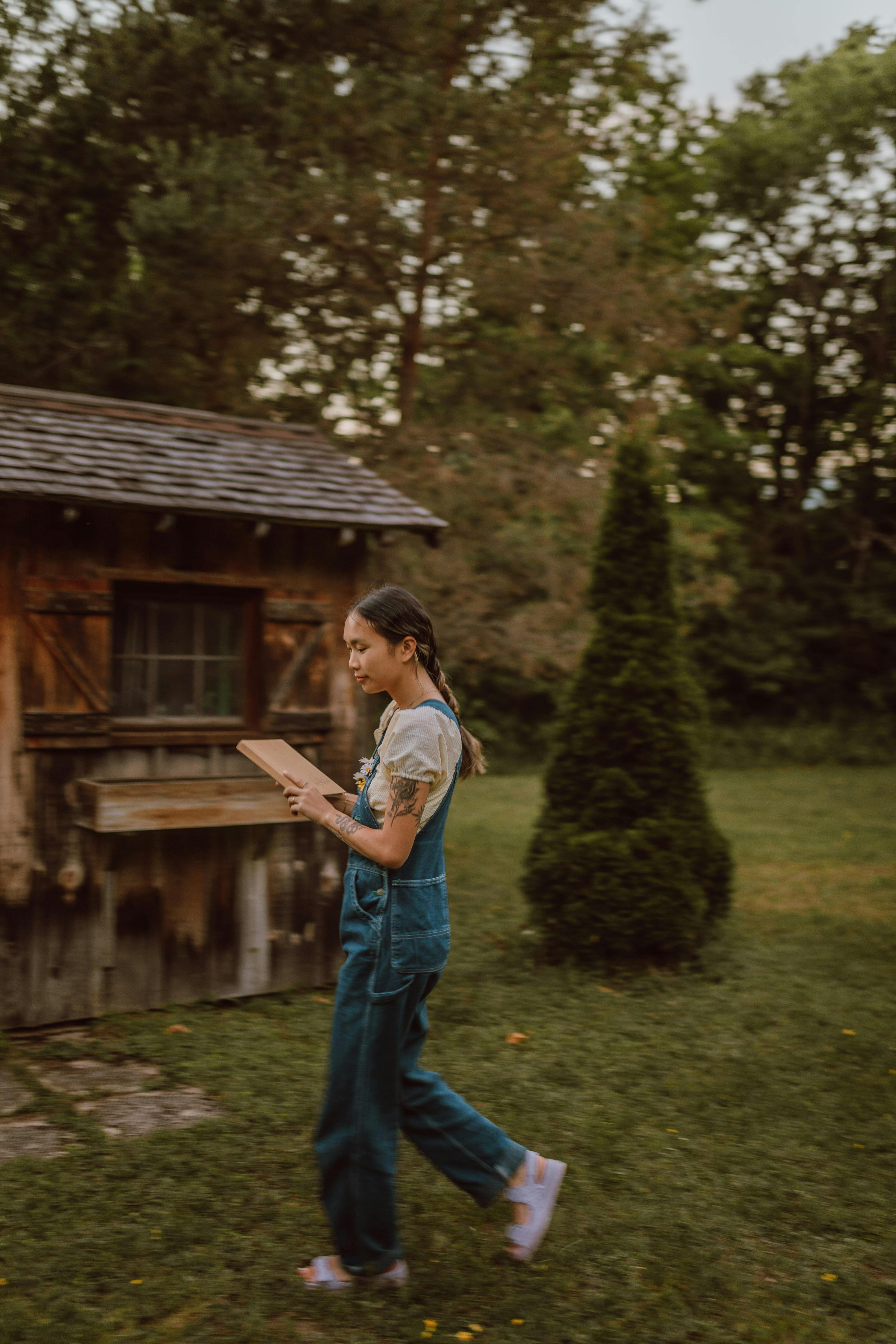 woman in denim overall walking on grass near barn and holding a book
