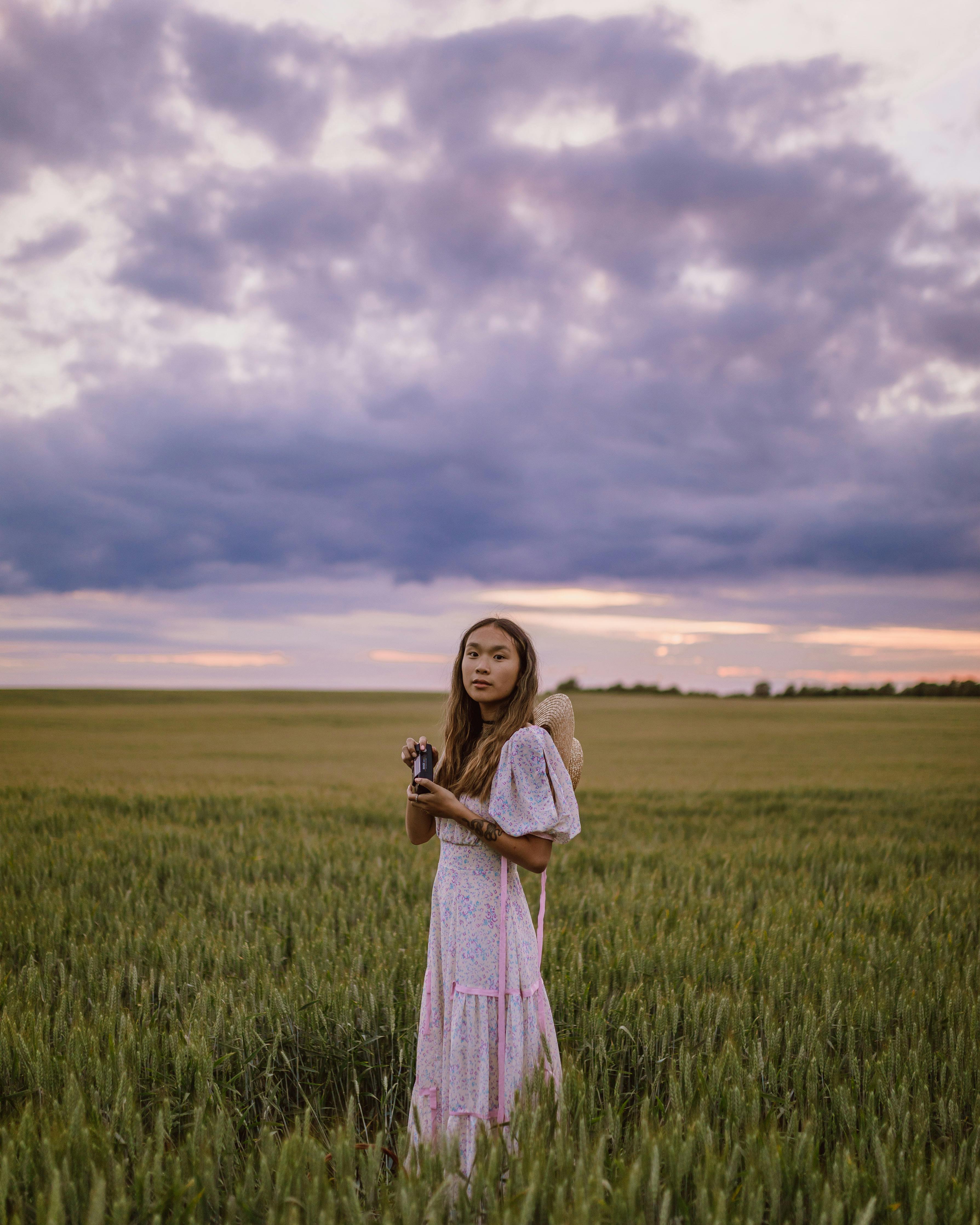 woman in white dress standing in green field and holding a camera during sunset