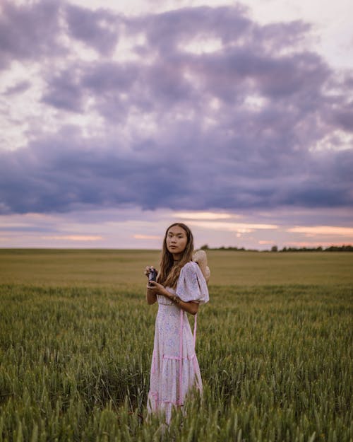 Woman in White Dress Standing in Green Field and Holding a Camera During Sunset