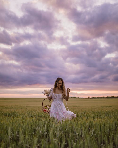 Young Woman in White Dress Walking in Green Field and Holding Basket with Flowers