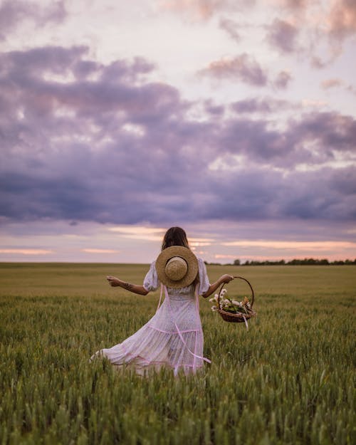 Free Woman in White Dress with Hat on Back and Basket in Hand Walking in Green Field Stock Photo