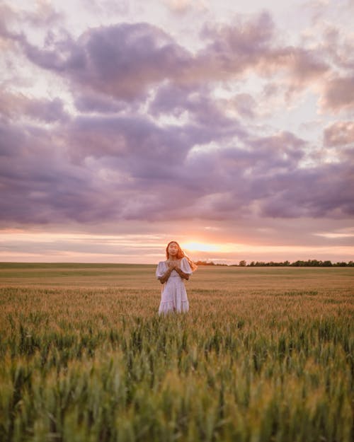 Woman in White Dress with Hands on Chest Standing in Field During Sunset
