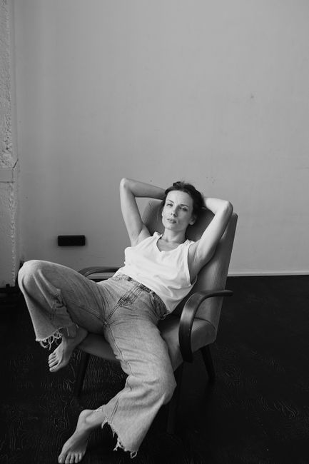 Woman wearing denim pants and white top, sitting relaxed on a chair in a black and white setting.