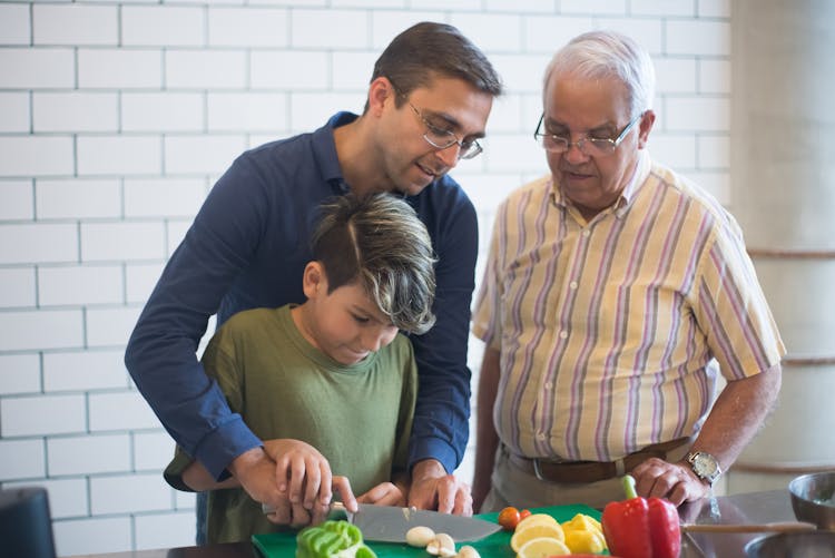 Elderly Man In Striped Shirt Standing Beside Man In Blue Shirt Who Is Teaching Boy Cutting Vegetables