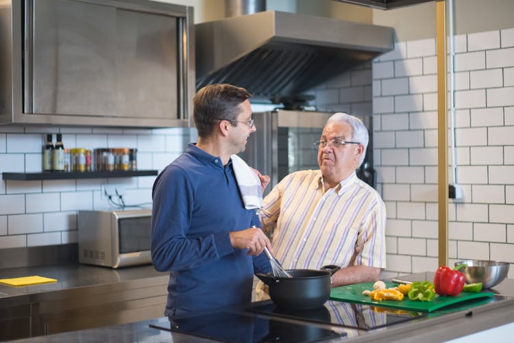 Elderly Man In Striped Shirt Talking To Man In Blue Shirt With Towel On Shoulder In The Kitchen