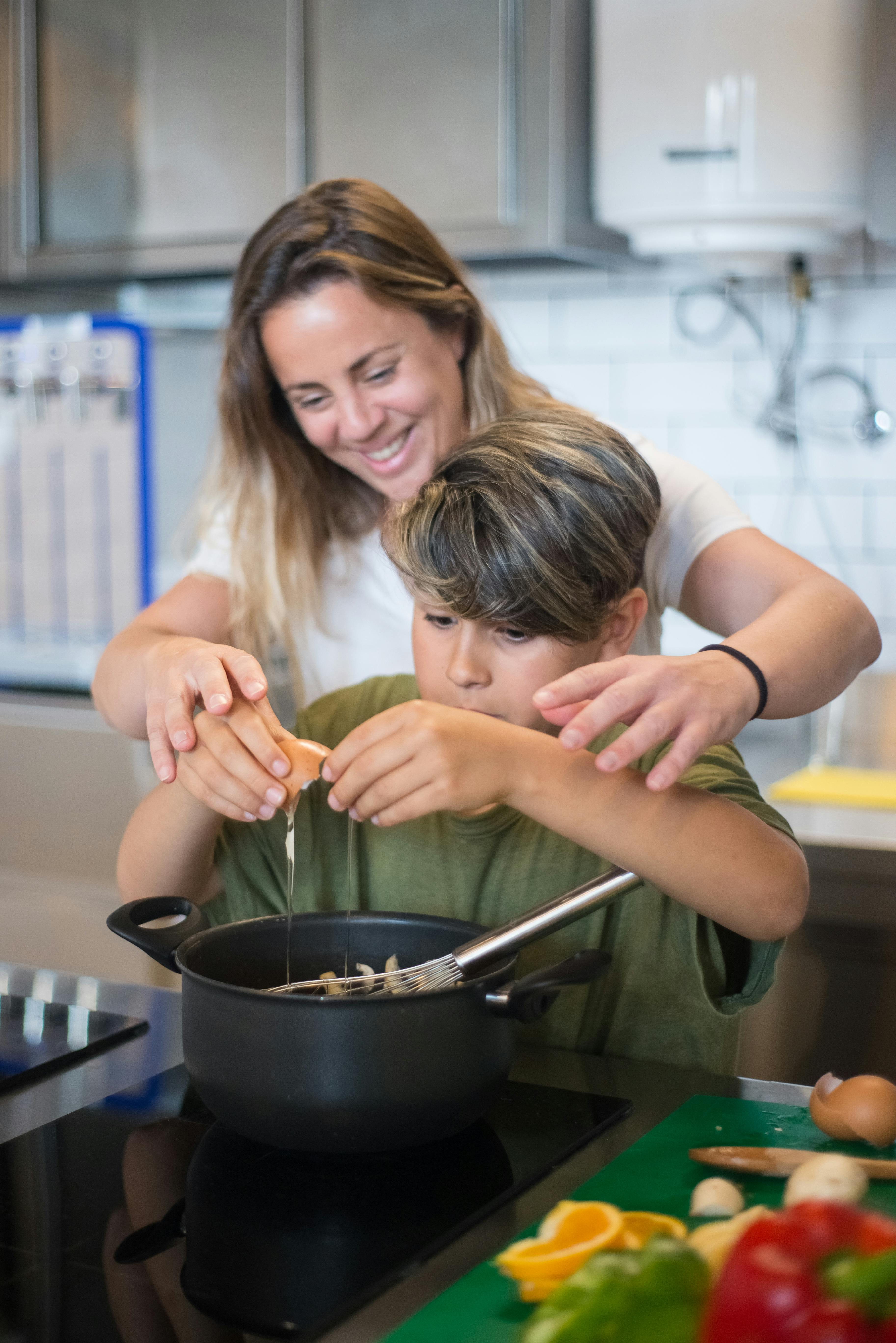 woman in white t shirt standing behind a boy breaking an egg into pan