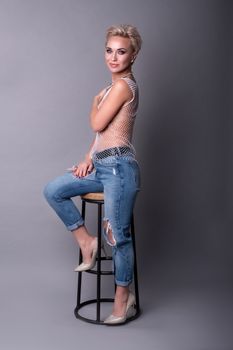 Woman with short blonde hair in trendy attire sitting on a stool against a grey backdrop.