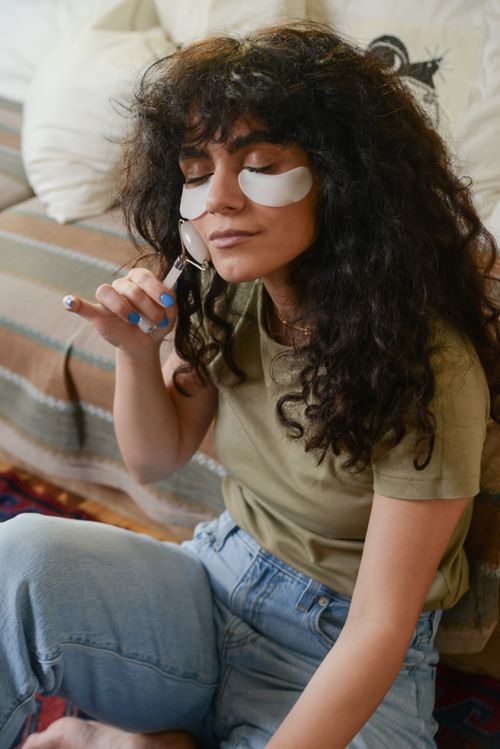 Free Woman with curly hair using a jade roller for skin care, wearing under-eye patches. Stock Photo