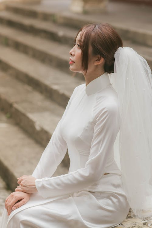 Free Woman in White Long Sleeve Dress with Veil on Her Hair Sitting on a Concrete Stairs while Looking Afar Stock Photo