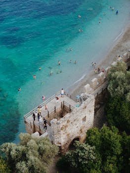 Drone view of Antalya's coastline with a historical tower and swimmers enjoying the turquoise waters.