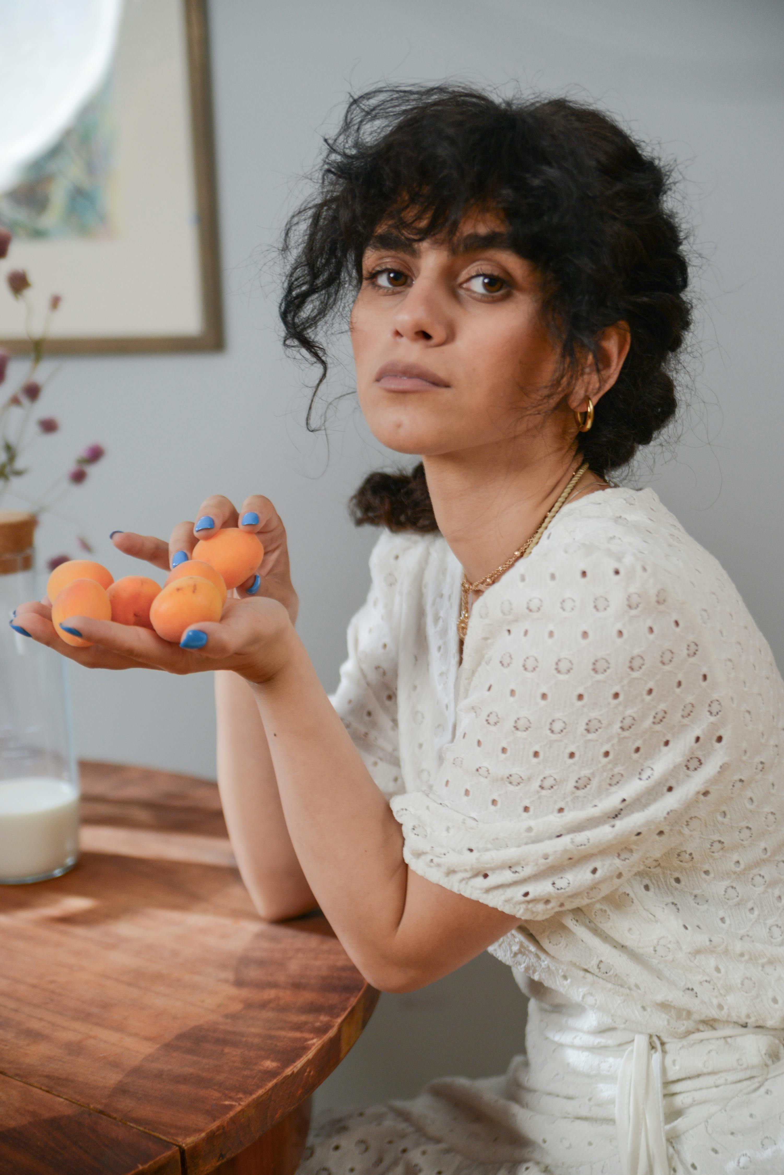 Woman in a white dress with apricots, sitting at a wooden table indoors.