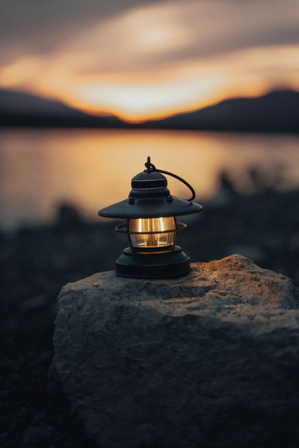 Warmly lit lantern resting on a rock by the water at sunset, creating a cozy atmosphere.