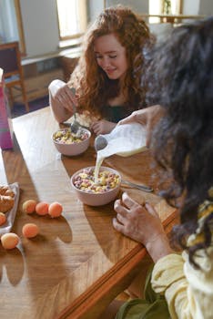 Two women enjoying a breakfast of cereal with milk in a sunlit kitchen.