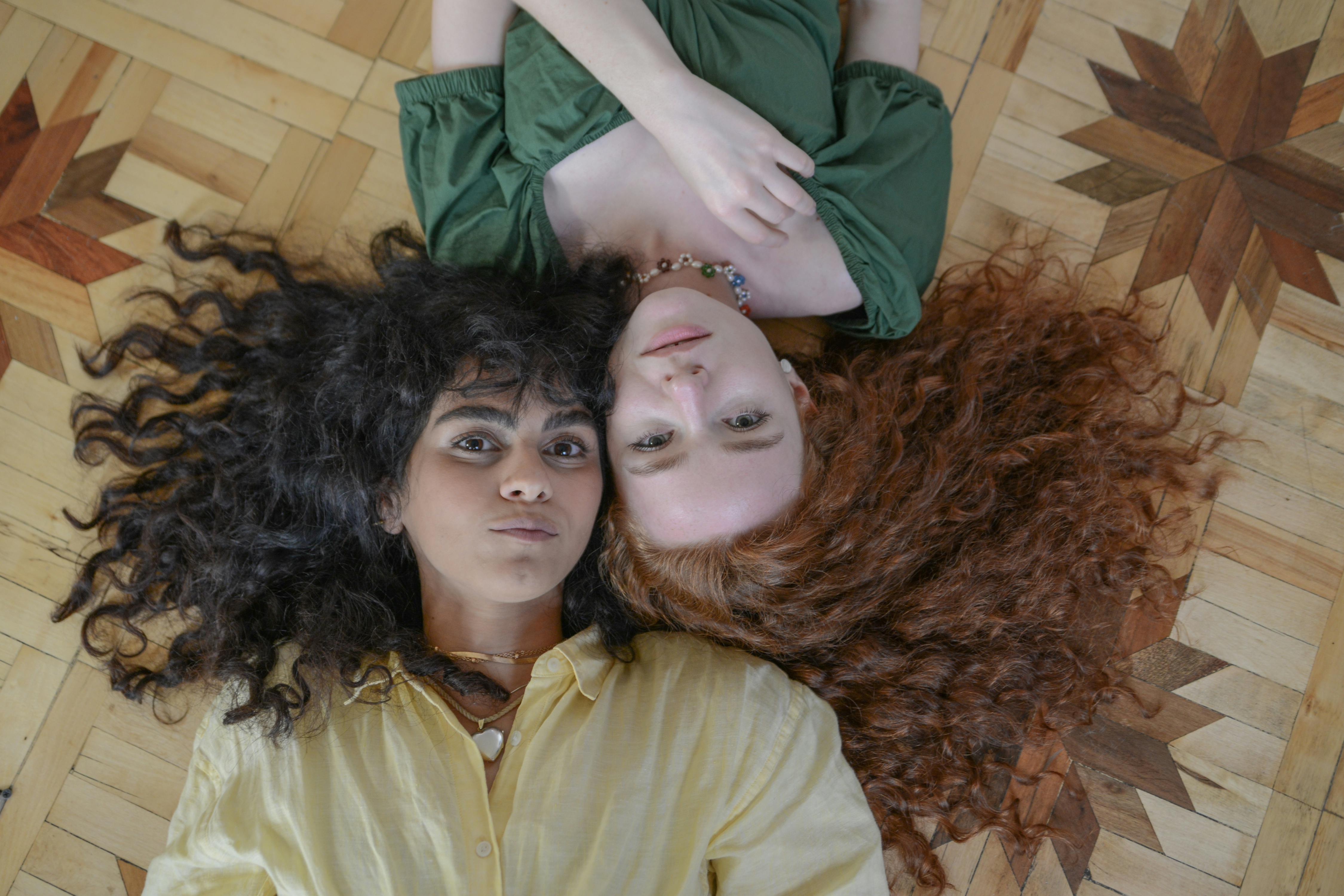 Two women with curly hair lying head to head on a patterned floor, showcasing friendship and style.