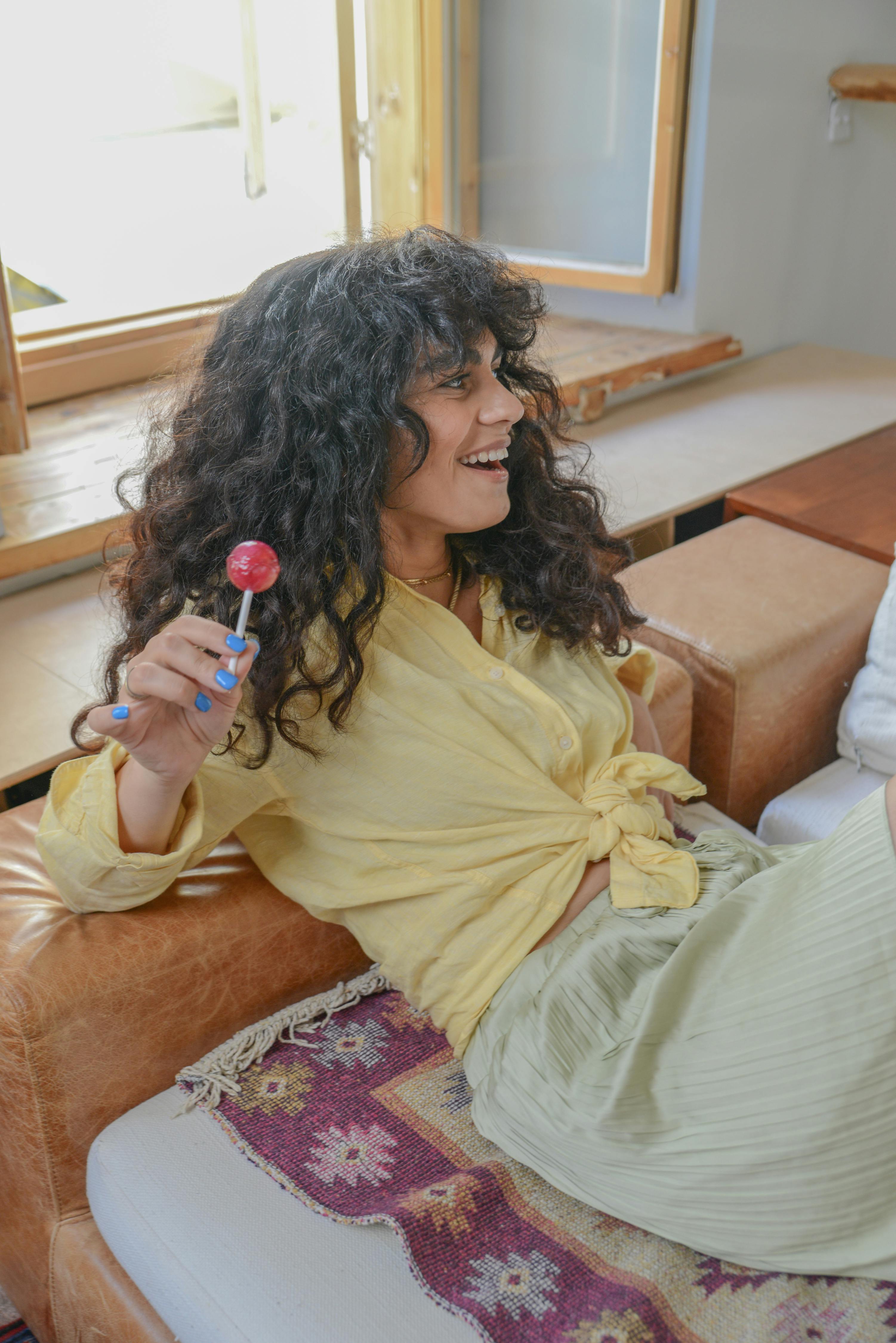 Cheerful woman with curly hair enjoying a lollipop while relaxing indoors.