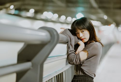 Free A contemplative young woman in long sleeves leans against a railing with a blurred urban background. Stock Photo