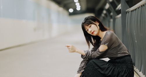 Free A young woman in a stylish outfit sits by a metal fence in a softly lit urban environment. Stock Photo