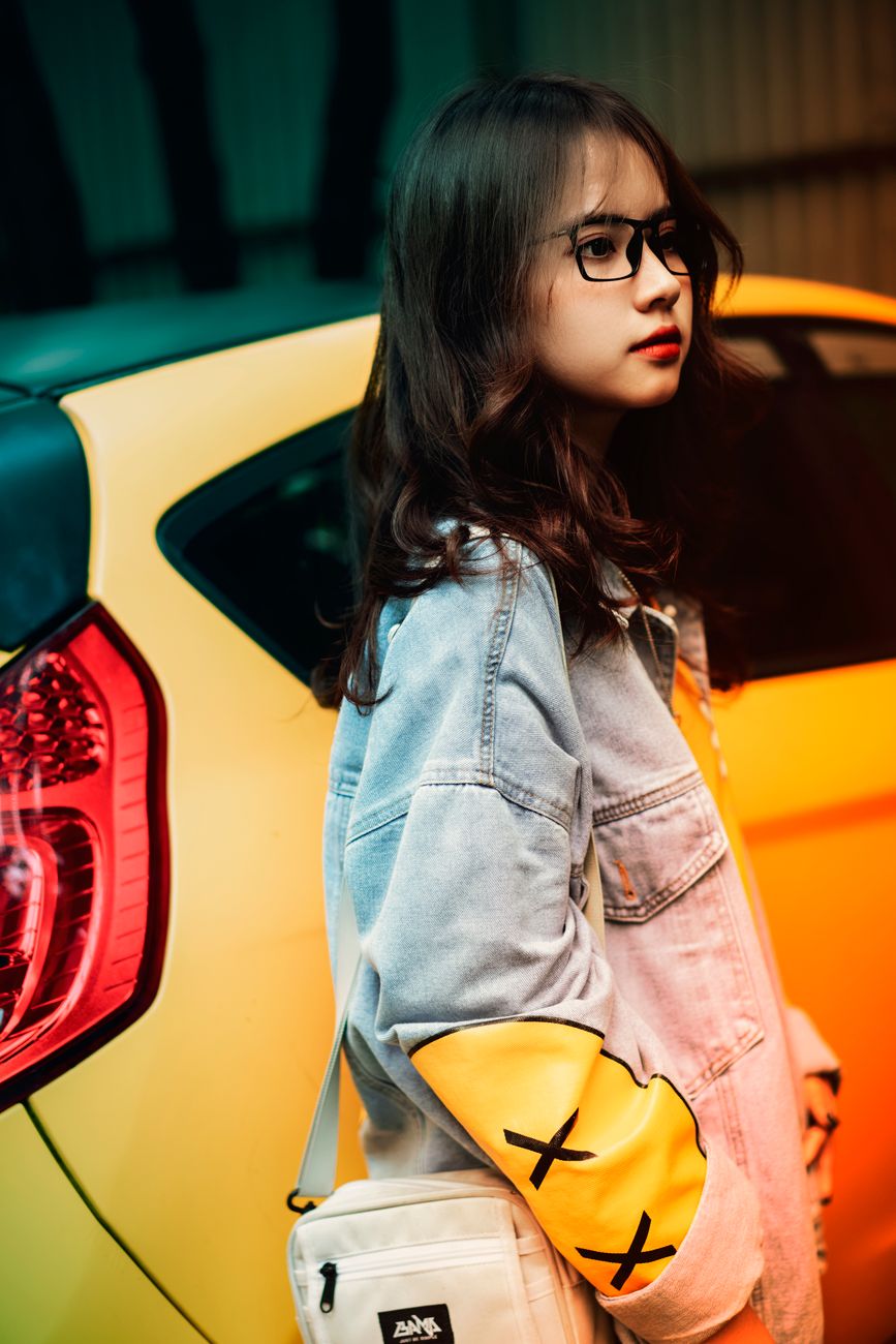 Fashionable young woman with eyeglasses and denim jacket stands confidently by a stylish yellow car.