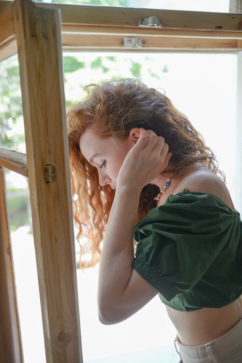 Free Side view of a young woman standing by an open window, bathed in natural light. Stock Photo