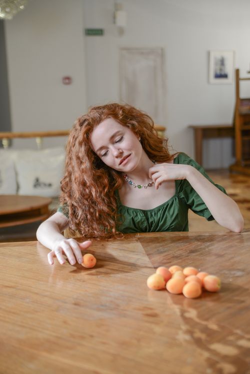 Free A red-haired woman in a green blouse sits at a table with apricots, creating a serene indoor ambiance. Stock Photo