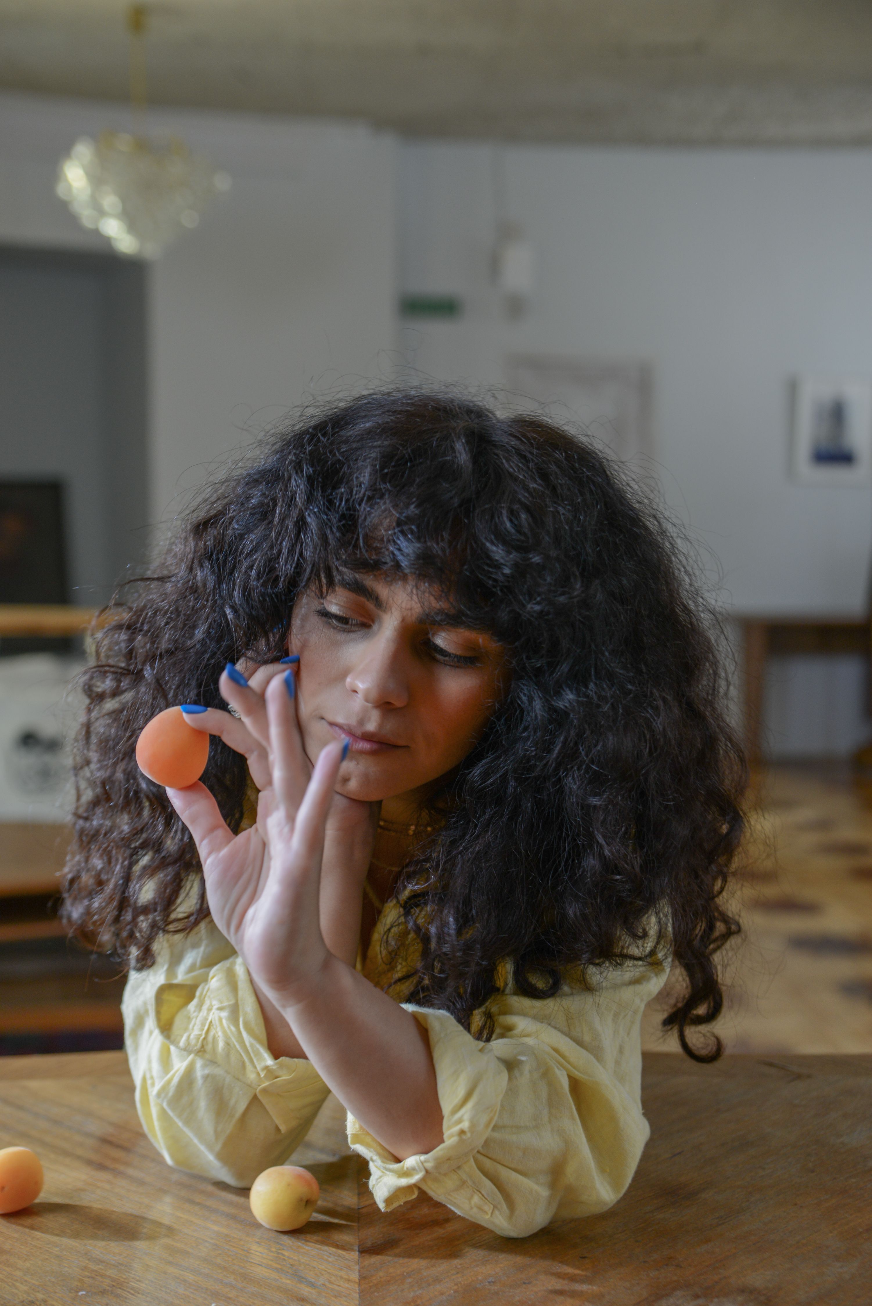 A woman in a yellow shirt thoughtfully holding an apricot inside a room.