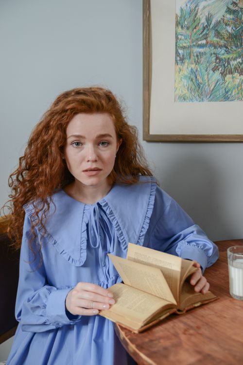 Free Red-haired woman in a blue dress reading a book indoors, creating a serene atmosphere. Stock Photo