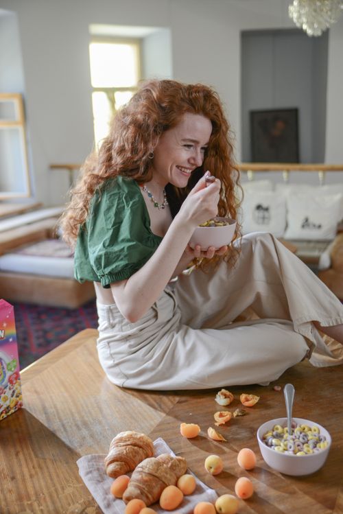 Free Redheaded woman happily enjoying a breakfast bowl at home with pastries and fruits. Stock Photo