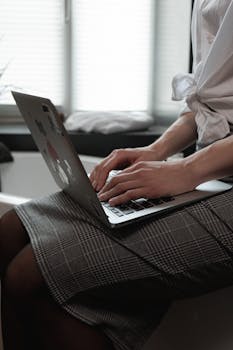 A person typing on a laptop while sitting indoors near a window, wearing a skirt.