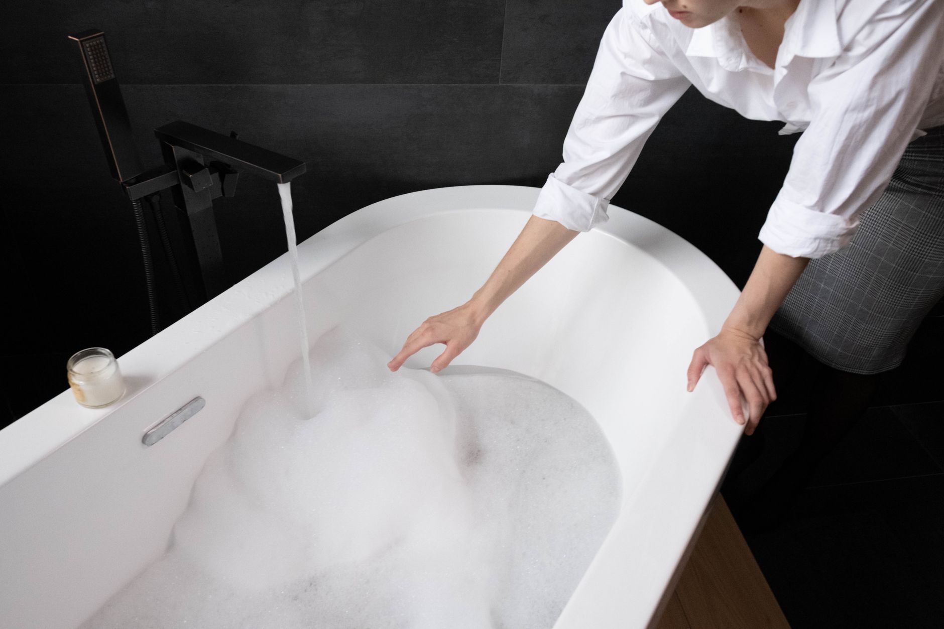 A woman in a bathroom runs water into a foam-filled bathtub, creating a relaxing atmosphere with a candle nearby.