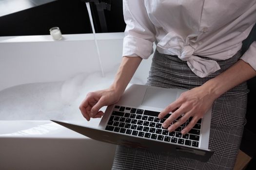 Adult woman working on a laptop while sitting by a bathtub filled with bubbles, combining work and relaxation.