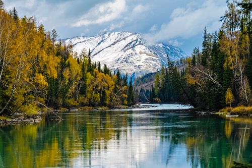 Free Tranquil river flows through autumn forest with snow-covered mountains in the background. Stock Photo