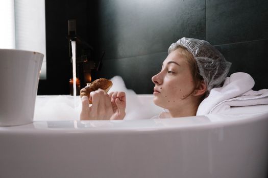 A woman relaxes in a bubble bath, wearing a shower cap, and enjoying a pastry.
