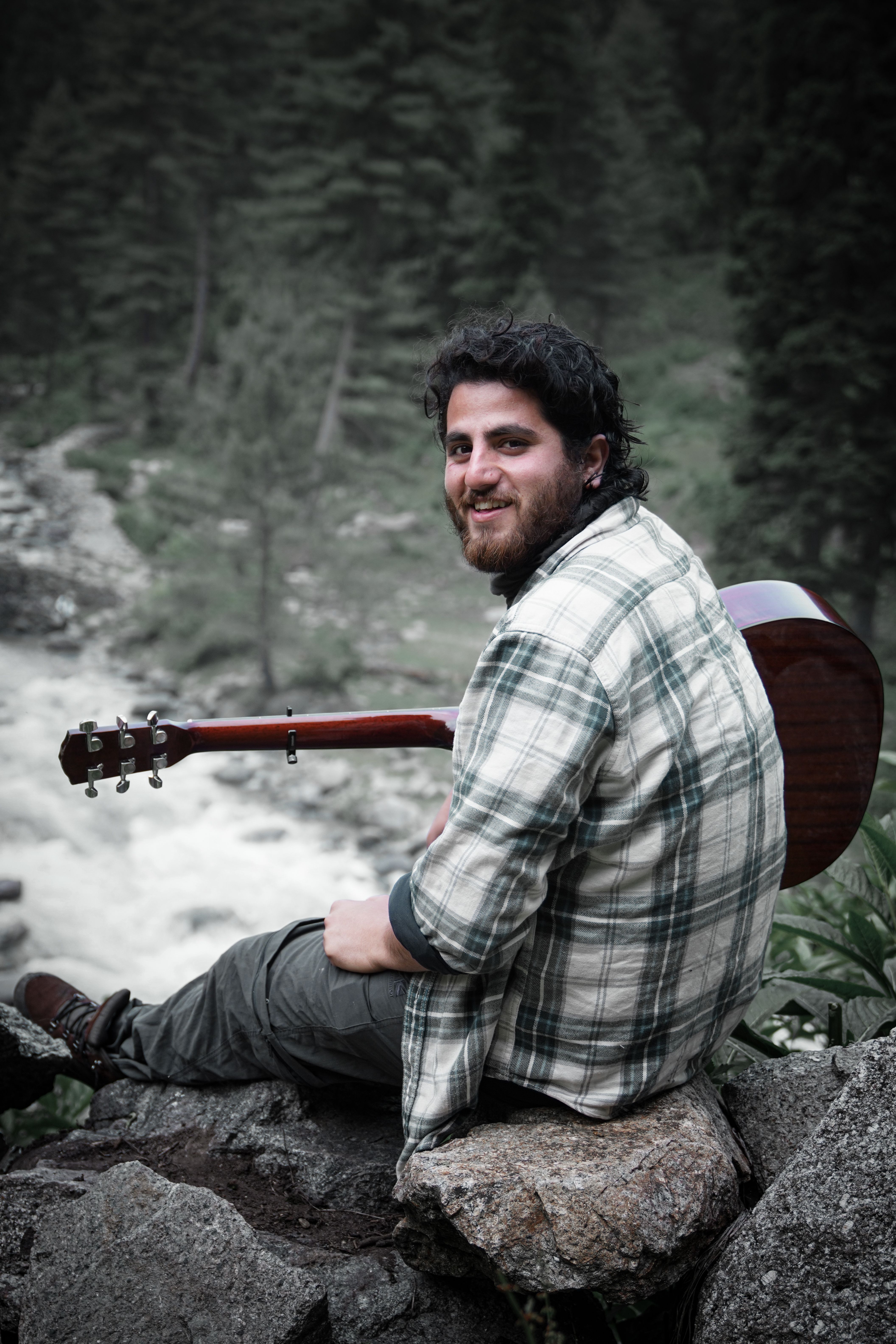 A man sits on rocks with a guitar, surrounded by lush greenery in Wangat.