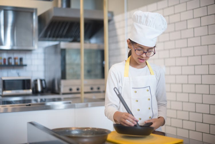 A Young Girl In White Long Sleeves Cooking While Holding A Pan